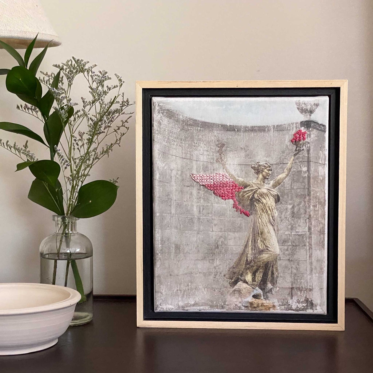 Framed 8"x10" art with pink embroidered wings and bowl of the Spirit of Life photo on a shelf with white clay bowl, and glass vase with green foliage and white flowers.