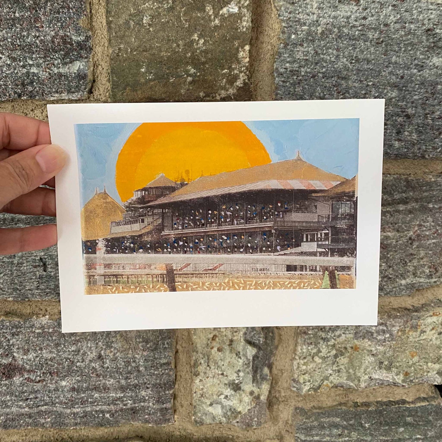 Hand holding 5"x7" fine art print of the historic Saratoga Racecourse - mixed media with photography, blue, gold, and yellow acrylic paint and embroidery.